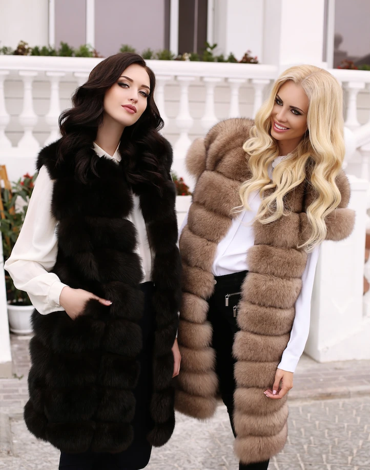 Two women wearing long faux fur vests in black and taupe
